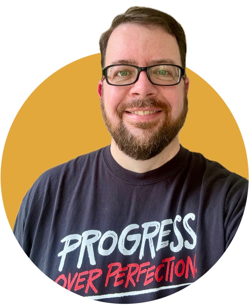 Matt Vanderzalm, a tall white person wearing glasses, smiles at the camera. Matt is wearing a black t-shirt that says Progress over Perfection.